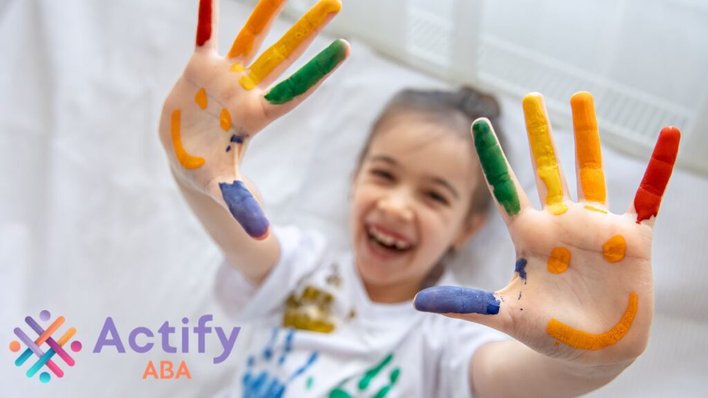 A young girl holds out both hands toward the camera, palms facing outwards, painted with a smiley face and colorful designs, symbolizing creativity and support for autism advocacy.