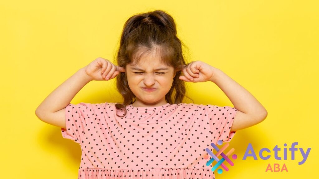 A young girl in a checkered dress covers her ears in a bright yellow room, showing discomfort from sound sensitivity often experienced by children with autism.