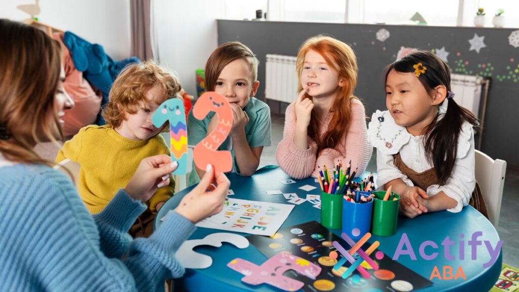 A group of children, including those with autism, sit together in a therapy room, listening as an instructor holds up colorful numbers for an interactive lesson.